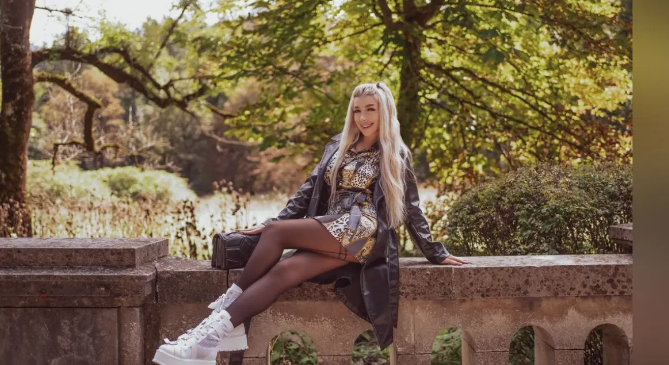 Das Cam Girl Sierra in einem herbstlichen Park, stilvoll mit Lederjacke und Sneakers.